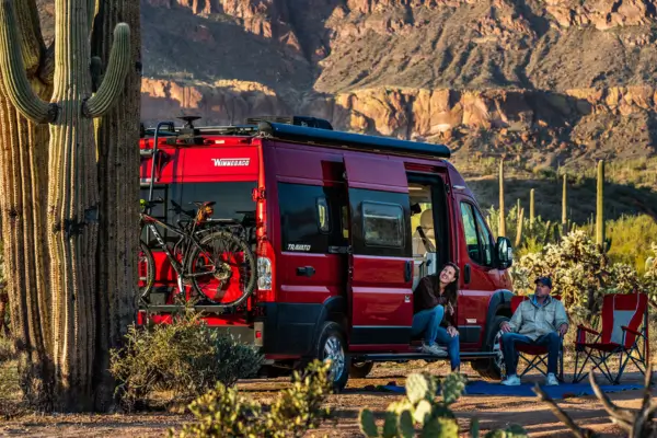 Mini motorhome vermelho Winnebago® estacionado no deserto com casal sentado ao lado aproveitando a vista entre cactos e montanhas — ideal para viagens com modelos compactos.